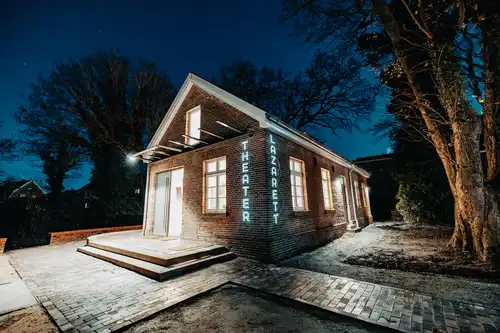 Weitwinkel: Ein von innen leuchtendes Backsteinhaus im Dunkeln mit dem Logo `Theater Lazarett`