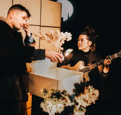 Ein Mann mit Miniaturbaum in der Hand und eine Frau mit E-Gitarre schauen sich an.